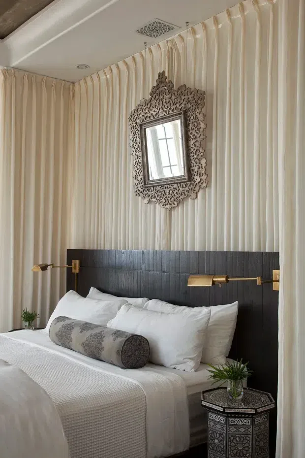 A refined bedroom featuring full-height cream drapery covering the walls, a dark textured built-in headboard, white bedding, brass wall-mounted reading lights, and an ornate carved mirror above the bed. A patterned decorative side table and small plants add detail. The palette is warm neutral with dark and brass accents, creating a luxurious, intimate atmosphere.
