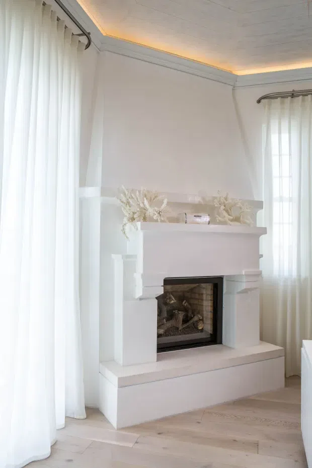 Bright, elegant interior corner centered on a substantial white-painted fireplace surround with a dark firebox. Full-height sheer curtains frame tall windows on two sides, while a pale timber floor, white walls, decorative cornices, and recessed warm cove lighting create a refined, airy setting. Dried botanical arrangements and small decor sit on the mantel.