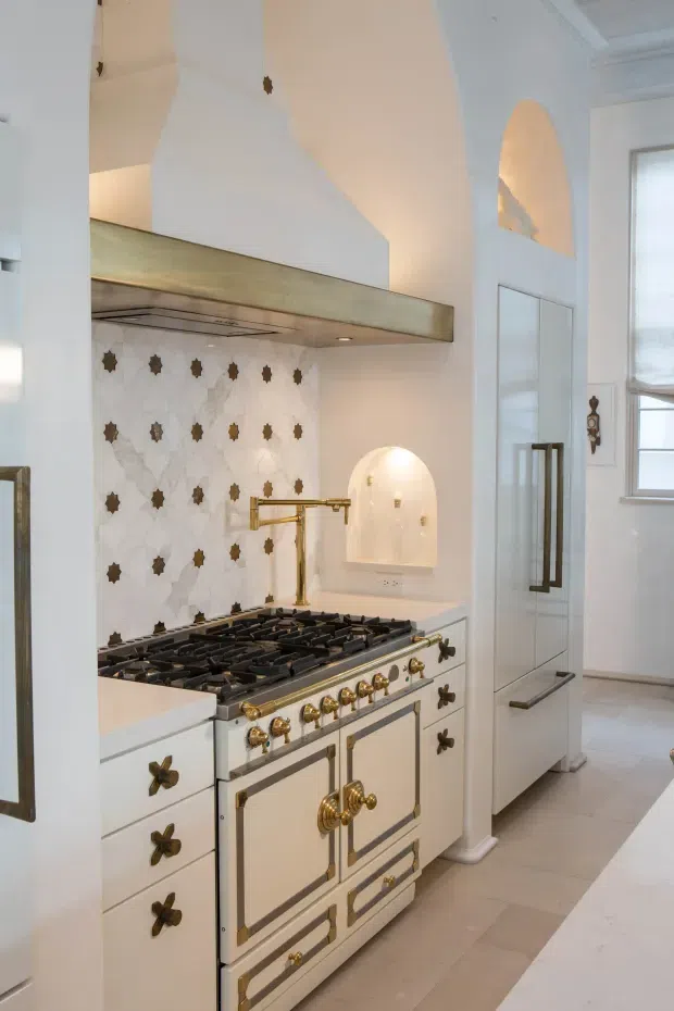 A refined traditional kitchen centered on a large white range cooker with ornate brass hardware. The kitchen features white fixed cabinetry, a decorative white-and-grey patterned tile splashback with repeated dark metallic motifs, a brass pot filler, pale stone or tile flooring, and a substantial pale metallic rangehood canopy. Arched recessed niches, tall cabinetry, classic trim, and a bright window reinforce the heritage-inspired design.
