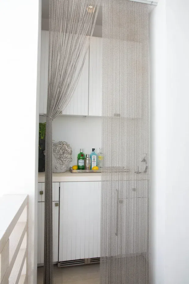 Bright, narrow utility area with white vertically panelled built-in cabinetry, a pale benchtop, and a decorative hanging chain curtain partially screening the space. Bottles and a small tray sit on the counter, with a white railing visible in the foreground and light neutral tiled flooring.