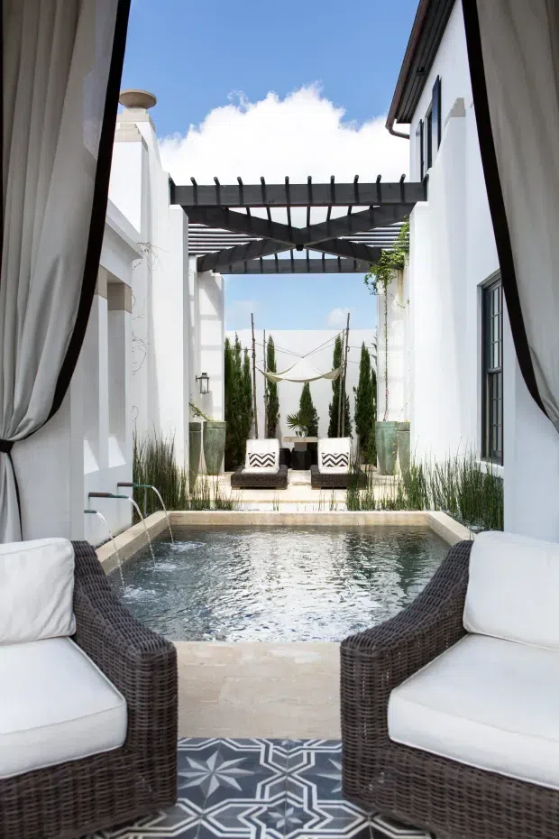 A bright courtyard-style outdoor living space viewed from between white curtains, with two wicker lounge chairs in the foreground overlooking a rectangular plunge pool. White rendered walls, dark timber pergola beams, tall narrow planting, decorative planters, and a pair of outdoor lounge chairs create a refined resort-like setting.