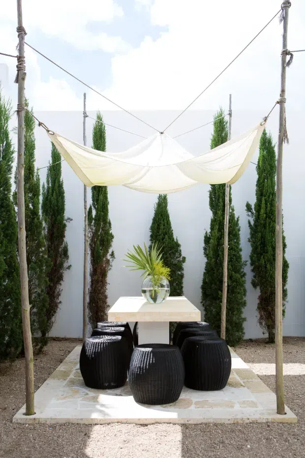 A small outdoor courtyard dining area framed by tall slender evergreen trees and four rustic timber poles supporting a stretched white fabric canopy. A pale stone-paved platform holds a square light-coloured dining table, a round glass vase with tropical foliage, and several glossy black rounded stools. The setting is bright and minimal, with gravel ground and a pale rendered wall.