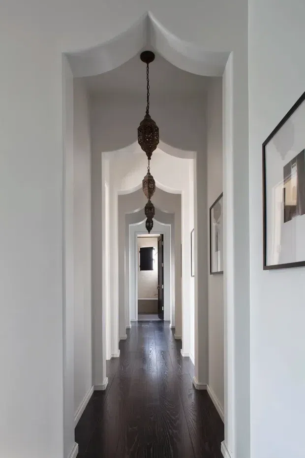 A long, narrow interior hallway with repeated white arched ceiling openings and door surrounds, dark timber flooring, white walls and skirting, framed artwork, and a series of ornate hanging lantern pendants creating a strong perspective toward the far doorway.