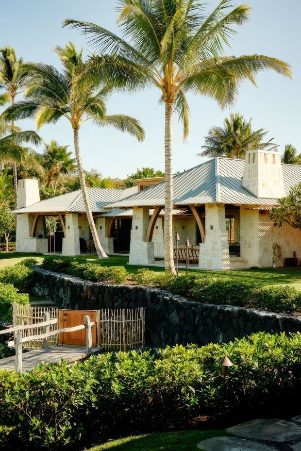 A tropical single-storey house surrounded by tall coconut palms and lush landscaping. The building features pale stone walls, a light grey metal hipped roof with prominent roof planes and chimney-like masonry elements, deep eaves, timber-framed openings, and curved timber structural supports. In the foreground are a dark volcanic-stone retaining wall, a timber pedestrian gate, and manicured hedges.