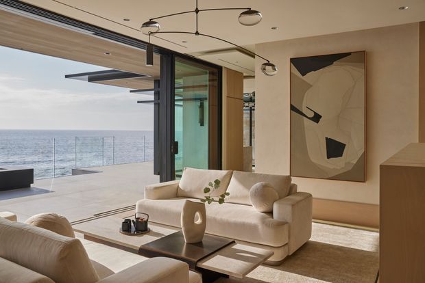 Refined contemporary living room opening onto a spacious ocean-facing terrace through extensive floor-to-ceiling glazing. The room features a cream boucle-style sofa and armchairs, low dark coffee tables, warm timber joinery, neutral plaster walls, a large abstract artwork, and sculptural suspended lighting. The bright, airy interior has a strong indoor-outdoor connection and expansive sea views.