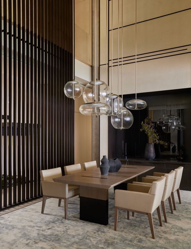 A refined contemporary dining room with a large rectangular timber dining table, upholstered cream dining chairs, and a dramatic cluster of suspended glass globe pendant lights. Full-height dark vertical timber slats form a feature wall beside warm beige wall panelling, while a dark reflective wall and sculptural ceramic vessels add depth. The room has a high ceiling and a muted patterned rug beneath the dining setting.