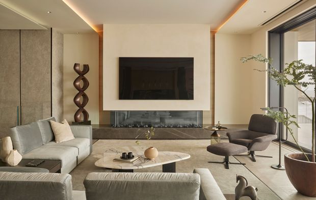 A refined contemporary living room with a large built-in fireplace and television feature wall, low-profile sectional seating, sculptural coffee table, designer lounge chair, and tall indoor plant beside full-height glazing. The space uses warm neutral finishes, layered concealed lighting, dark stone around the fireplace, and a spacious open layout.
