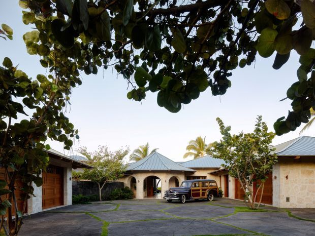A low-rise tropical courtyard compound viewed beneath a canopy of broad-leaf trees. The courtyard is paved with irregular dark stone slabs and includes arched openings, timber garage or entry doors, pale stone walls, and multiple low hipped metal roofs. Two vintage vehicles are parked centrally, with palms and lush planting visible around the buildings.