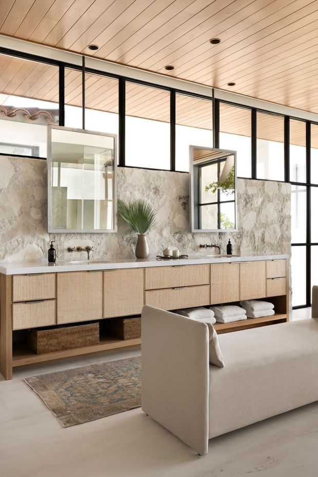 A bright, spacious bathroom with a long double vanity in woven-texture light timber cabinetry, white stone benchtop, open shelving, and two wall-mounted basins with brushed metal tapware. The walls feature a rough natural stone finish, while a timber-lined ceiling and full-height black-framed glazing create a warm contemporary feel. Two framed mirrors, neutral furnishings, folded towels, baskets, and a sculptural vase complete the refined spa-like interior.