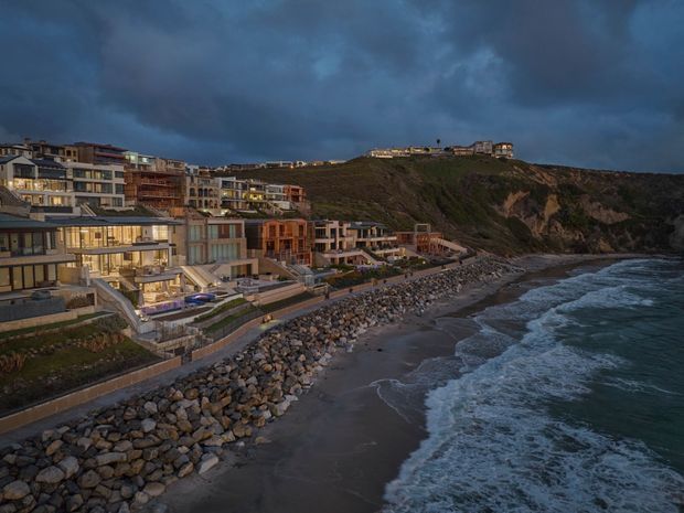 Aerial coastal view of a dense, upscale multi-storey residential development built along a rugged ocean shoreline. The buildings step up the hillside and feature extensive glazing, balconies, terraces, and warm contemporary lighting. A rocky seawall and narrow beach meet the surf below, with dramatic overcast clouds creating a moody atmosphere.