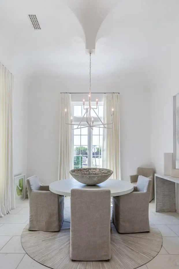 Bright, elegant dining room with a round white dining table surrounded by upholstered slipcovered chairs. The room features tall curtain-dressed glazing, a sculptural multi-arm chandelier, white curved ceiling detailing, pale tiled flooring, and a large round woven rug with a stone bowl centerpiece. A textured console or cabinet is visible to the right.