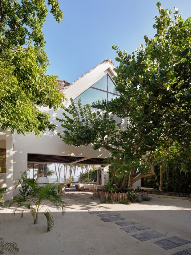 A bright tropical resort-style building courtyard framed by dense greenery. The white rendered structure has a steep gabled roof with a thatched or textured roof edge, a large triangular glazed window, and an open shaded ground-level living area with timber ceiling slats. Pale paving, stepping stones, palms, and planted garden beds lead toward a glimpse of water and palms beyond.