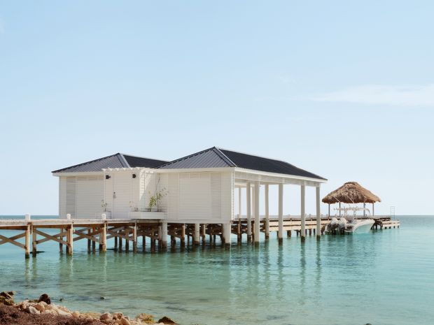 A white coastal building raised on stilts over clear turquoise water, with multiple dark pitched roof sections, louvred or shuttered wall panels, open verandah-like structures, and a timber pier leading to a thatched-roof gazebo. The scene is a bright tropical waterfront setting viewed from the shoreline.