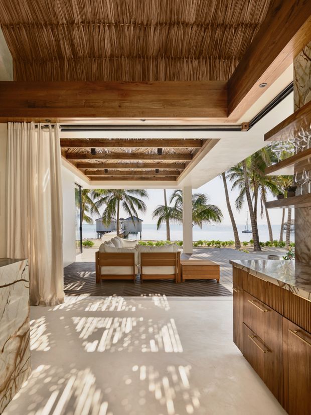 An open-plan tropical coastal living and entertaining area opening directly onto a beachfront terrace. The space features a woven reed or thatched ceiling, exposed timber beams, pale plaster walls, full-height open glazing with cream curtains, warm timber cabinetry, and a stone-clad counter with open shelving. Lounge seating faces a sandy beach, palm trees, and the ocean beyond, creating a bright indoor-outdoor connection.