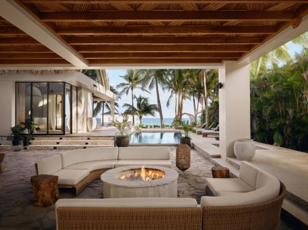 A luxurious open-air covered outdoor lounge facing a tropical beachfront setting. The space features a broad timber-beamed ceiling, white rendered columns and walls, pale stone paving, curved cream upholstered seating, woven rattan furniture, and a round built-in fire pit. A swimming pool, palm trees, and ocean are visible beyond the lounge, with glazed interior spaces to the side.