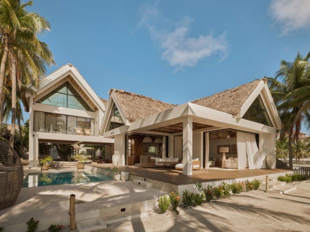 Exterior view of a tropical resort-style residence with multiple steep gabled roofs, prominent thatched roofing, open-air living spaces, pale structural framing, expansive glazing, timber decking, and a swimming pool surrounded by palms and landscaped planting.