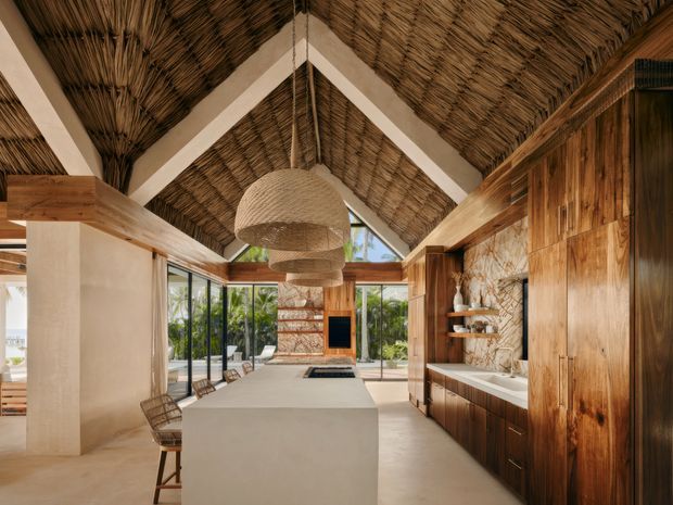 A spacious tropical contemporary kitchen and living area with a dramatic high pitched ceiling lined in woven natural thatch or reed, exposed white structural beams, extensive timber cabinetry and wall panelling, and a large pale stone island. Woven pendant lights hang above the island, while full-height glazing opens the room to lush greenery. A fireplace or media feature is visible at the far end, and open shelving with decorative objects lines the kitchen wall.
