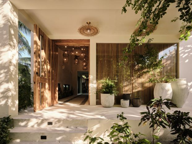 A warm, sunlit residential entry courtyard with a covered porch, broad concrete steps, timber pivot door, vertical timber screening, and large sculptural planters. The open doorway reveals a dimly lit interior hall with decorative pendant lighting. Tropical planting and dappled shadows create a relaxed indoor-outdoor atmosphere.