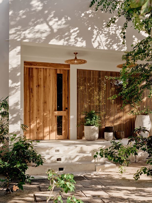 A warm, contemporary residential entrance viewed from outside. A tall natural timber door with a narrow dark glazed insert sits within a white rendered facade, beside a vertical timber slat screen. A woven pendant light hangs beneath the recessed entry canopy, with potted plants and leafy garden vegetation framing the steps. Dappled sunlight creates strong organic shadows across the walls and paving.