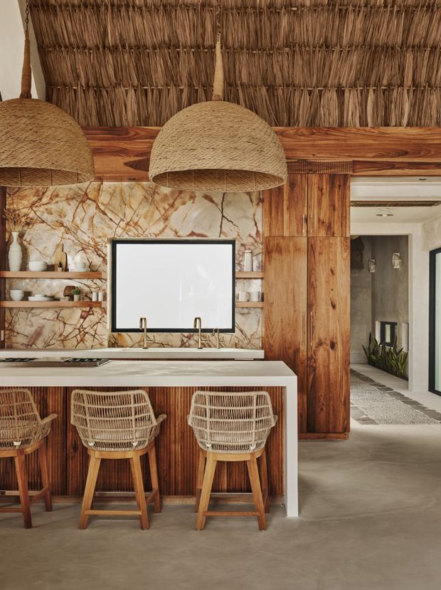 A warm, textural kitchen interior featuring a large island with a pale stone benchtop, timber and woven cabinetry, three woven rattan bar stools, and oversized natural-fibre pendant lights. The back wall combines dramatic veined stone, open timber shelving, and a timber-framed serving or window opening. A woven reed ceiling and timber structural detailing create a strongly organic, rustic aesthetic, while a hallway is visible beyond the kitchen.