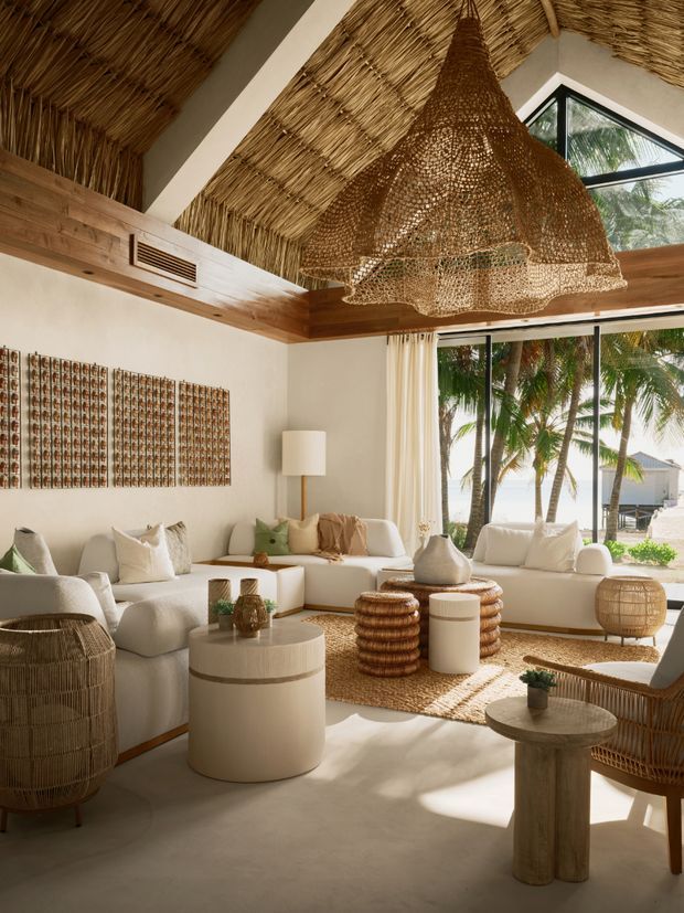 A bright, airy living room with a high pitched ceiling clad in woven reed or thatch, exposed timber beams and a large sculptural woven pendant light. The room features a cream sectional sofa, white occasional tables, woven ottomans and baskets, rattan seating, natural fibre rug, and warm timber detailing. Large glazed openings provide views toward palm trees and a sandy coastal setting.