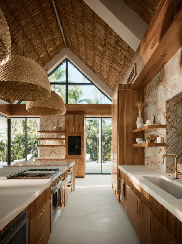 A warm, high-ceilinged contemporary kitchen with extensive dark timber cabinetry, pale stone benchtops, and dramatic veined stone splashback and wall surfaces. The room features a large central island with integrated cooktop, open shelving, a built-in oven, and a generous sink run. Woven pendant lights and a woven reed-lined vaulted ceiling add natural texture, while expansive black-framed glazing and a triangular clerestory window bring in abundant daylight and views of tropical vegetation.