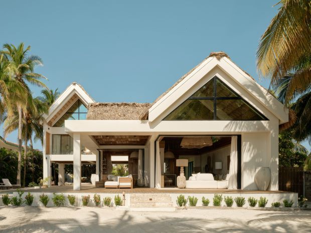 A tropical resort-style residence with a white rendered facade, steep triangular gables, broad open glazing, and thatched roof sections. The open ground-level living space is framed by white columns and opens onto a pale paved terrace with low planting. Palm trees surround the building, creating a bright coastal atmosphere.