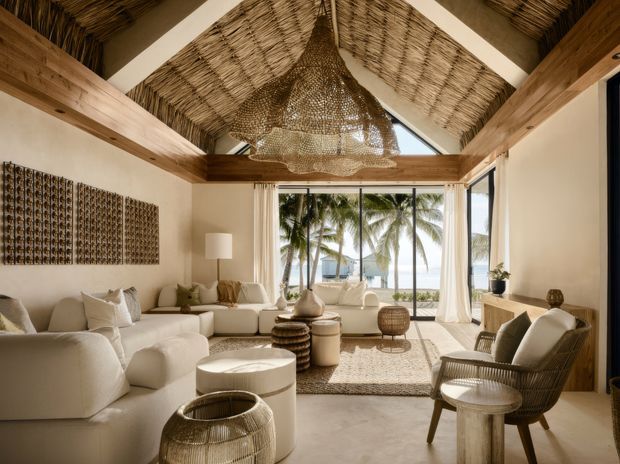 A bright, refined coastal living room with a large cream sectional, woven occasional tables, natural-fibre rug, rattan lounge chairs, and a sculptural woven pendant. Exposed timber beams frame a steeply pitched, woven-textured ceiling, while full-height glazing opens to a tropical beachfront view with palm trees and ocean beyond.