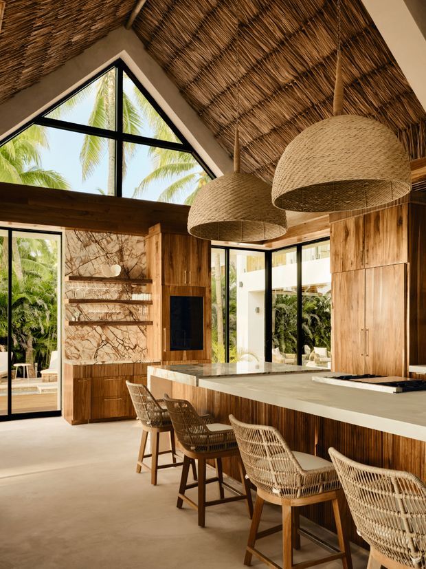 A luxurious tropical kitchen with a large island, woven bar stools, extensive warm timber cabinetry, and a marble-patterned feature wall. The room has a dramatic thatched or reed-lined vaulted ceiling, oversized woven pendant lights, black-framed floor-to-ceiling glazing, and views of palms and lush greenery outside.