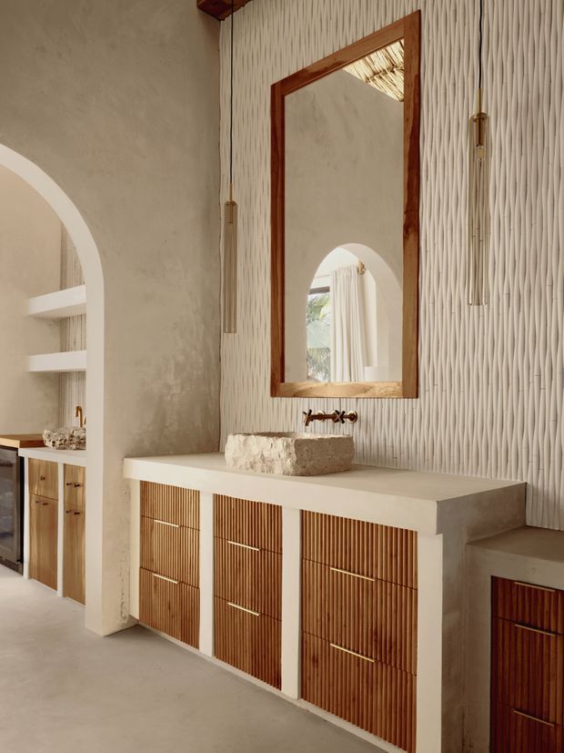 A warm, sculptural bathroom vanity area with a floating-style stone basin set on a pale plastered counter, ribbed timber cabinetry, and a large wood-framed mirror. The walls combine smooth textured plaster with a vertically patterned three-dimensional surface, while arched openings reveal additional shelving and cabinetry. Brass tapware and pendant details add subtle contrast. The overall composition is earthy, refined, and Mediterranean-inspired.