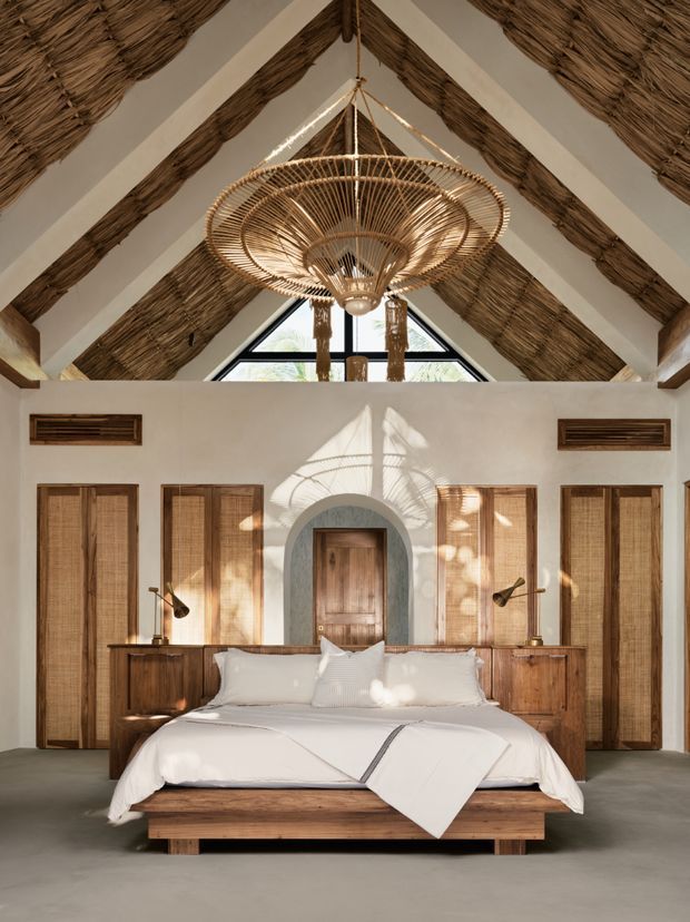A warm, refined bedroom with a centered bed beneath a dramatic pitched ceiling. The ceiling features exposed timber beams and woven natural-fibre thatching, complemented by a large woven pendant light. White plaster walls, timber louvred doors, built-in timber cabinetry, an arched doorway, and a dark neutral floor create a calm, earthy retreat. Strong sunlight casts patterned shadows across the room.