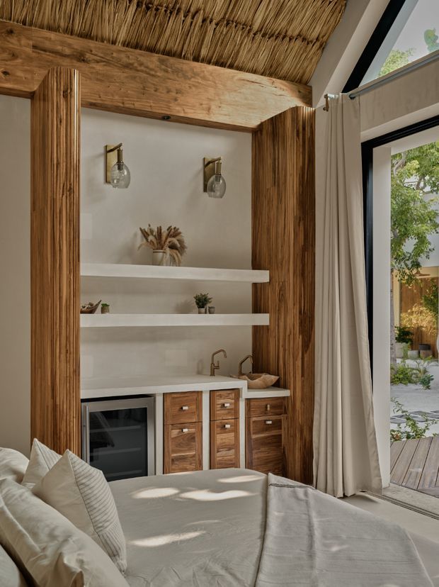 A warm, textural bedroom interior featuring a built-in timber wet bar or beverage station with a compact refrigerator, drawers, sink and open shelving. Vertical timber panelling frames the joinery, while a rustic woven reed ceiling adds strong natural texture. A large upholstered bed occupies the foreground beside full-height black-framed glazing and linen curtains, opening toward a planted outdoor courtyard.