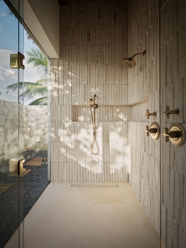 A luxurious open walk-in shower with pale vertically textured wall tiles, a matching light stone or tiled floor, and brushed brass shower controls and fixtures. A frameless glass panel opens toward a small pebble-floored outdoor courtyard, with tropical palm foliage visible beyond a low stone wall. Natural dappled sunlight creates a warm, resort-like atmosphere.