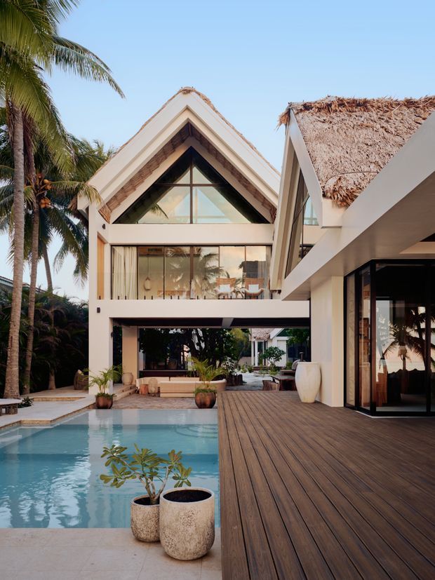 A tropical luxury residence surrounds a swimming pool and broad timber deck. The prominently visible building has crisp white rendered walls, steep gabled forms, large triangular glazing, and a thatched roof section, with open-plan indoor-outdoor connections and palm landscaping.