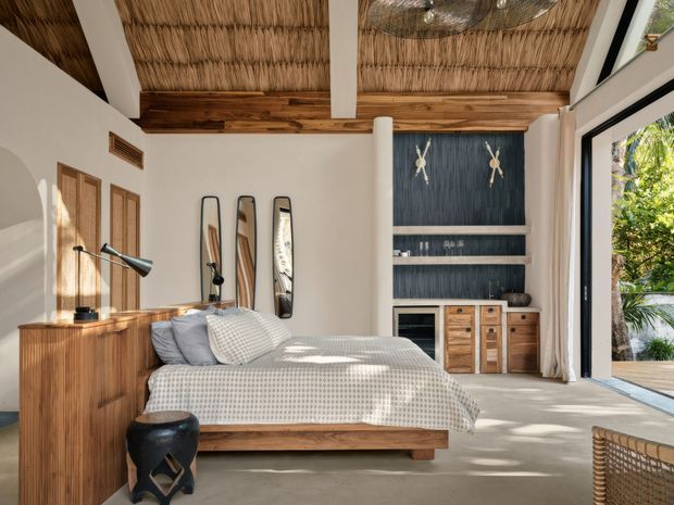 A sunlit bedroom with a low timber bed, textured neutral bedding, timber bedside cabinetry, a matching timber dresser, surfboard-shaped wall decor, and a dark built-in shelving and media unit. The room features a thatched or woven ceiling with exposed timber beams, white plaster walls, concrete flooring, and expansive black-framed glazing opening toward lush greenery. The overall atmosphere is warm, relaxed, and resort-like.