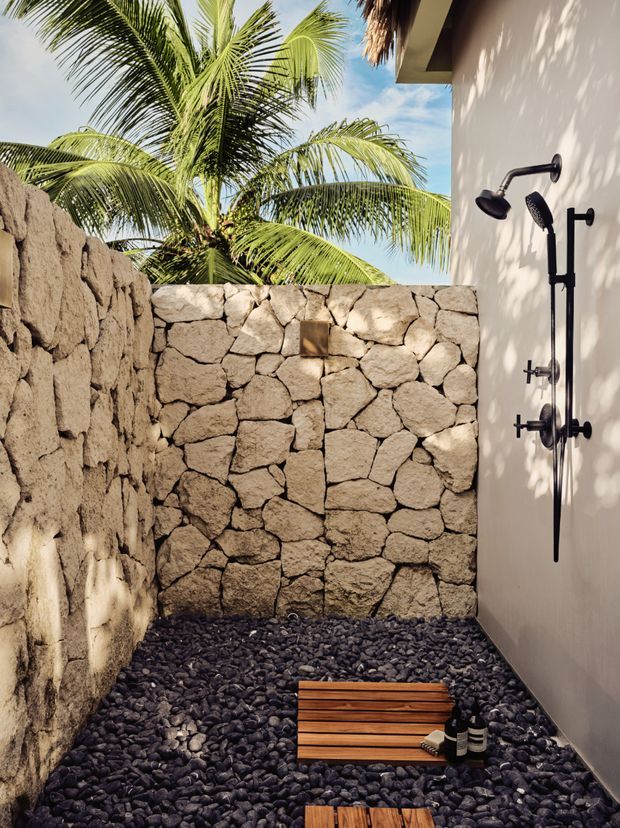 An open-air outdoor shower courtyard with a black multi-head shower fixture mounted on a smooth light wall. Rough natural stone walls enclose the space, while dark rounded pebble flooring and small timber slatted shower platforms create a spa-like setting. Tropical palm foliage and bright sky are visible above the wall, with strong natural sunlight and shadows.