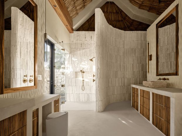 A sculptural, spa-like bathroom with a walk-in shower, freestanding toilet, dual vanity areas, and large framed mirrors. The room features white vertically textured wall tiles, warm timber-framed joinery with slatted cabinet fronts, brass fixtures, a smooth pale floor, and an exposed rustic woven or thatched ceiling with timber beams. Natural light enters through the glazed shower opening and door, creating a warm, organic atmosphere.