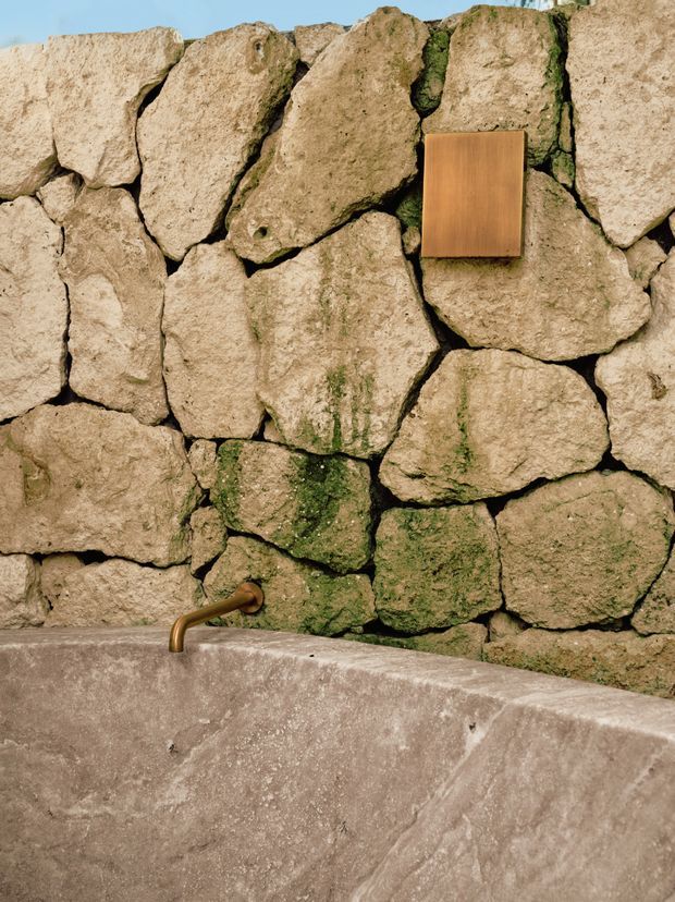 Close-up of a rustic outdoor or spa-like bathing area featuring a rough natural stone wall with moss-filled joints, a weathered concrete or stone basin, and a small brass wall-mounted tap. A warm timber-toned rectangular wall plate or control is mounted above the basin. The palette is earthy and tactile, with strong natural textures and minimal detailing.