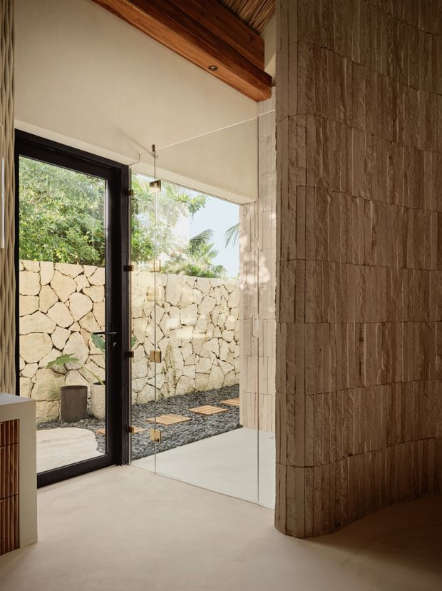 A warm, minimalist interior passage or bathroom-like space with a full-height frameless glass panel and dark-framed glazed door opening onto a private stone-walled courtyard. The room features a smooth pale floor and walls, a textured stacked-stone feature wall with a rounded transition, exposed timber ceiling beams, and a small built-in timber-fronted unit. Natural light, tropical vegetation, and a gravel courtyard create an organic indoor–outdoor connection.