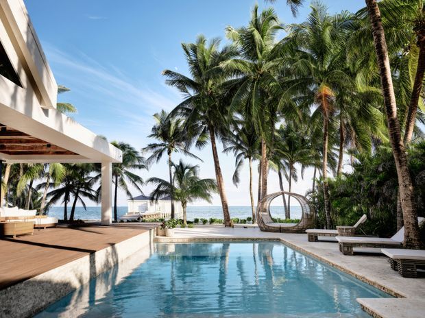 A bright tropical coastal resort-style outdoor space with a rectangular swimming pool beside a timber deck and covered open-air lounge. Tall coconut palms, dense greenery, ocean views, white modern architecture, sun loungers, and a hanging circular chair create a relaxed beachfront setting.