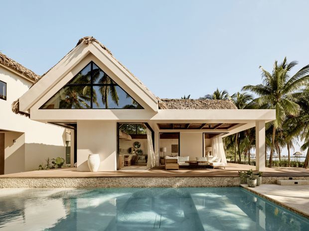A tropical resort-style house viewed from the pool, featuring a steep triangular gable with extensive black-framed glazing, open-plan covered living areas, cream stucco walls, and thatched roofing. Palm trees and a bright blue sky surround the property, while the foreground is dominated by a reflective swimming pool.