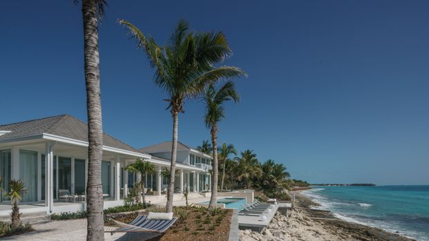 A low-rise coastal beachfront residence with pale rendered walls, extensive floor-to-ceiling glazing, hipped grey shingle roofs, and a series of connected wings stepping along the shoreline. Tall palm trees, a narrow pool, lounge chairs, rocky beach edges, turquoise water, and a clear deep-blue sky create a tropical resort-like setting.