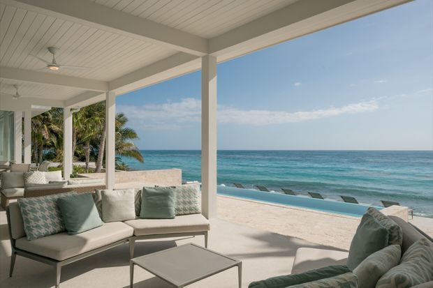 A bright, open-sided beachfront outdoor living area with a covered lounge, white painted posts and beams, a white paneled ceiling, ceiling fans, and pale upholstered seating around a low coffee table. The terrace overlooks a narrow pool and sandy beach with turquoise ocean beyond. The overall design is relaxed coastal and resort-like.