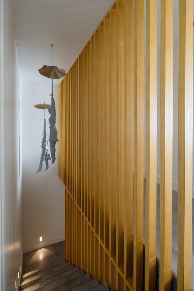 Interior view looking down a stairway beside a full-height vertical timber batten screen. The screen forms a warm golden feature wall and balustrade, with glimpses of the stair structure behind it. Dark stone or concrete steps descend toward a softly lit lower level. White walls and ceiling create a restrained backdrop, while sculptural hanging pendant lights add an organic decorative element.