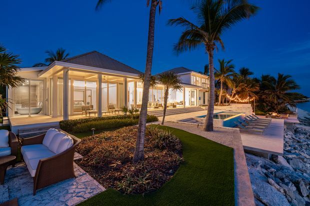 A luxurious low-rise beachfront resort-style residence at dusk, with illuminated pavilion-like rooms, extensive floor-to-ceiling glazing, pale rendered walls, dark hipped roofs, landscaped tropical gardens, outdoor lounge seating, a swimming pool, and a rocky shoreline beyond. The scene has a polished tropical coastal atmosphere with warm interior and landscape lighting.