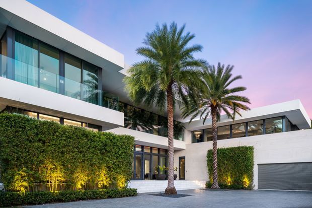 Exterior view of a large modern flat-roof residence with crisp white rendered walls, extensive dark-framed glazing, balconies, recessed lighting, and a wide integrated garage. Tall palm trees and carefully clipped green hedges frame a paved entry courtyard and driveway, creating a polished resort-like composition.