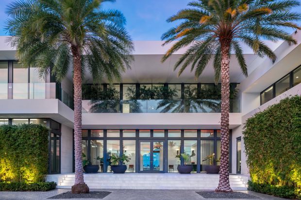Contemporary two-storey courtyard facade with broad glazed openings, dark-framed doors and windows, white rendered walls, and a covered upper-level terrace or walkway. Tall palm trees, clipped hedges, low planting, and illuminated landscaping frame the entrance, creating a resort-like modern exterior.