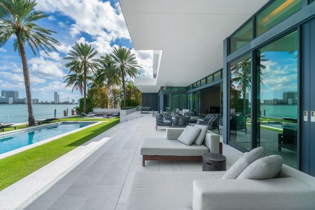 A bright contemporary waterfront residence viewed from a broad outdoor terrace. The terrace features pale large-format paving, multiple light upholstered lounge seating areas, floor-to-ceiling dark-framed glazing, a long cantilevered roof canopy, and a rectangular swimming pool beside manicured lawn and palm trees. High-rise buildings are visible across the water.