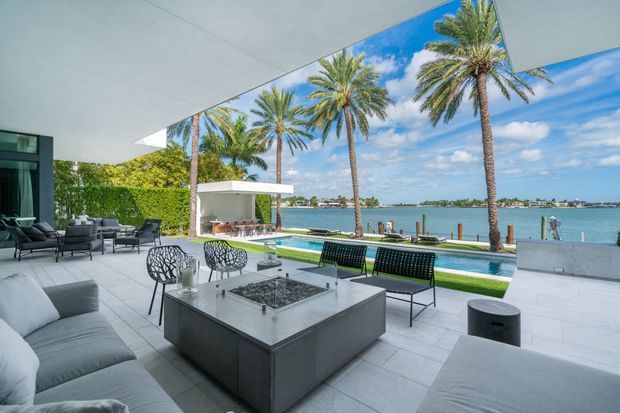 Contemporary waterfront outdoor living area with a covered terrace, pale tiled flooring, grey sectional seating, a central square fire table, and modern lounge furniture. A narrow swimming pool runs along the edge of the terrace, with palm trees and a calm body of water beyond. The space is open, bright, and resort-like, with clean architectural lines and extensive outdoor entertaining space.