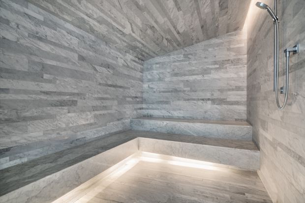 A luxurious walk-in shower or steam-room enclosure fully clad in pale grey veined stone or marble-look tile. The space features built-in tiered bench seating, a recessed illuminated floor or lower ledge, and minimalist chrome shower fixtures. The ceiling and walls are finished continuously in the same stone material, creating a calm, spa-like atmosphere.